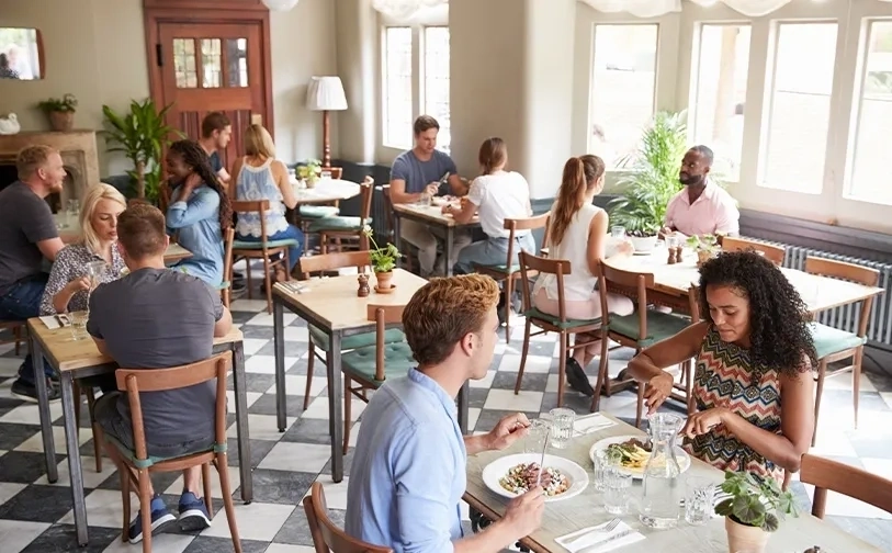 Ein helles, legeres Restaurant mit mehreren Tischen, an denen Menschen essen und sich unterhalten, ausgestattet mit Holzstühlen, schwarz-weißen Fliesen und großen Fenstern, die Tageslicht hereinlassen.