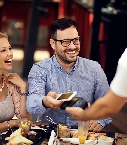 Person tätigt eine mobile Zahlung per Smartphone mit kontaktloser Technologie an einem Café-Tisch im Freien mit Getränken und Speisen.