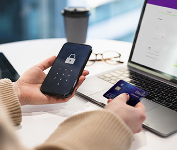 Person hält ein smartphone und eine kreditkarte vor einem laptop auf einem tisch mit einem kaffeebecher im hintergrund