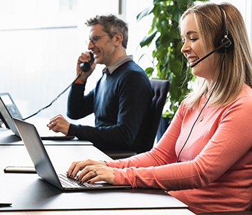 Zwei personen arbeiten an schreibtischen eine nutzt einen laptop mit headset und die andere spricht am telefon
