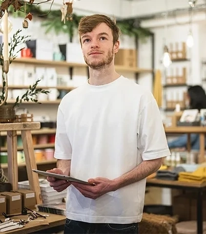 Eine Person in einem weißen Hemd hält ein Tablet in einem Geschäft mit Regalen voller Bücher, Kartons, Waren und dekorativen Pflanzen.